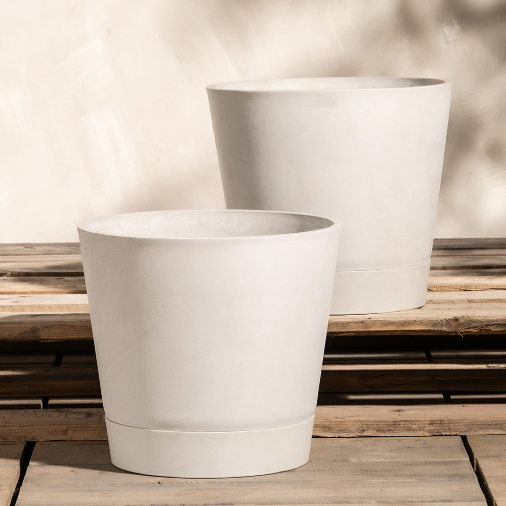 Zwei große, weiße Pflanzkübel-Sets aus Keramik (Venice, 2 x 30) stehen auf einem Holztisch vor einem neutralen Hintergrund. Die Töpfe sind zylindrisch und haben ein schlichtes, minimalistisches Design. Der Tisch hat ein verwittertes, rustikales Finish. Schatten werden auf die Hintergrundwand geworfen.