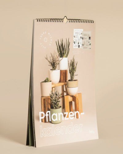 Der Pflanzenkalender ist ein stehender, spiralgebundener, immerwährender Kalender mit einem hellbeigen Hintergrund, der verschiedene getopfte Zimmerpflanzen auf Holzblöcken und den Titel "Pflanzenkalender" in großer weißer Schrift am unteren Rand zeigt.