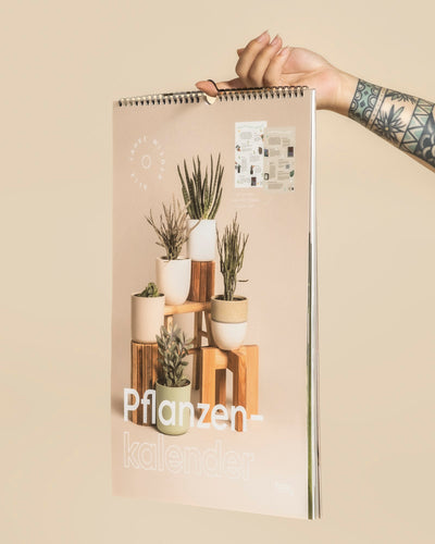 Eine tätowierte Hand hält einen spiralgebundenen Pflanzenkalender mit Zimmerpflanzen auf Holzständern vor einem beigen Hintergrund hoch. Auf dem Einband steht in großen weißen Buchstaben "Pflanzenkalender".