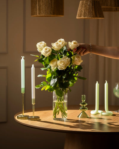 Eine Hand arrangiert einen Strauß Weiße Rosen in einer klaren Glasvase auf einem runden Holztisch, umgeben von grünen und weißen Kerzen in Haltern, bei warmer Beleuchtung, die Respekt und Aufrichtigkeit vermittelt.