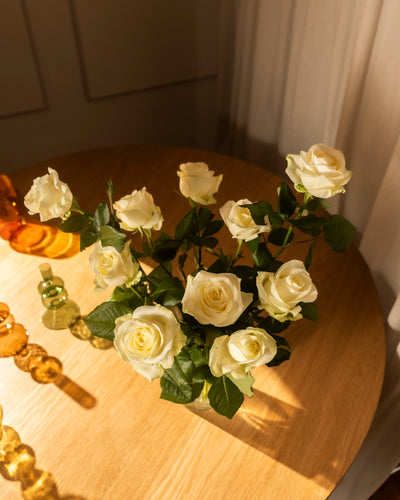 Ein Strauß Weißer Rosen mit grünen Blättern steht in einer Vase auf einem Holztisch, der in warmes Sonnenlicht getaucht ist, während dekorative Gläser auf der linken Seite Ehrlichkeit und Ruhe ausstrahlen.