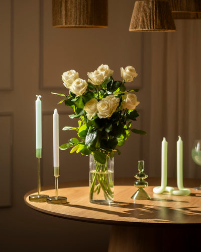 Ein Strauß Weißer Rosen in einer klaren Glasvase ruht auf einem Holztisch, umgeben von grünen und weißen Kerzen in modernen Haltern. Die warme Beleuchtung wirft weiche Schatten und schafft eine elegante, gemütliche Atmosphäre von Respekt und Ruhe.