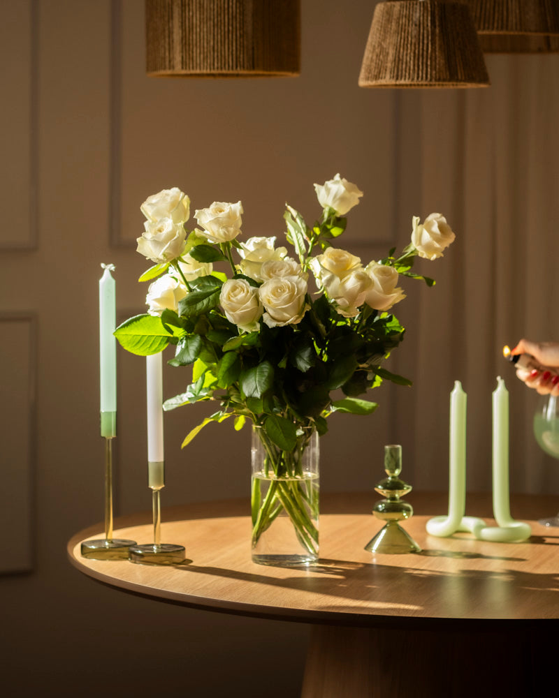 Ein Strauß Weißer Rosen in einer Glasvase ruht auf einem runden Holztisch mit grünen Kerzen. Eine Hand zündet eine Kerze an und füllt die Szene mit weichem Licht, das Ehrlichkeit und sanften Respekt widerspiegelt.