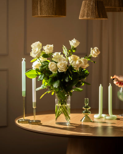 Ein Strauß Weißer Rosen in einer Glasvase ruht auf einem runden Holztisch mit grünen Kerzen. Eine Hand zündet eine Kerze an und füllt die Szene mit weichem Licht, das Ehrlichkeit und sanften Respekt widerspiegelt.