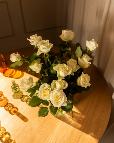 Der Weiße Rosenstrauß mit grünen Blättern steht in einer Vase auf einem runden Holztisch, umgeben von bernsteinfarbenem und grünem Glasdekor. Das Sonnenlicht unterstreicht die Szene und ruft Ehrlichkeit und Ruhe hervor.