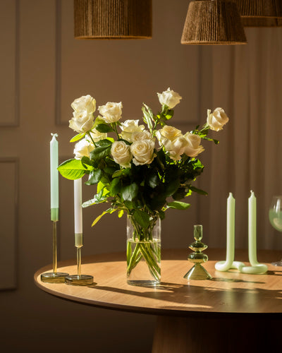 Eine Weiße-Rose-Vase steht auf einem runden Holztisch, umgeben von mintgrünen und weißen Kerzen in Glas- und Messinghaltern. Das warme Licht schafft eine gemütliche Atmosphäre, die Ehrlichkeit und Respekt widerspiegelt.
