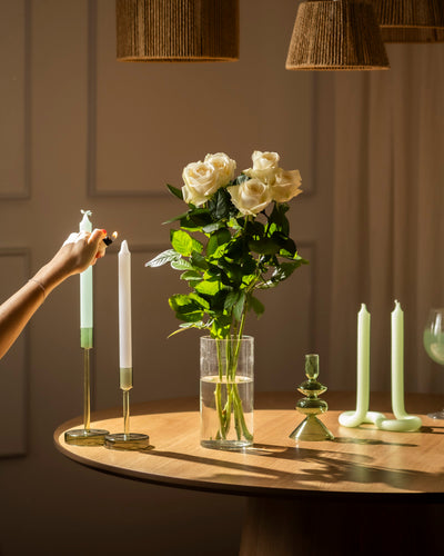 Eine Hand zündet eine weiße Spitzkerze auf einem runden Holztisch an, der mit Weißen Rosen in einer Glasvase, grünen und weißen Kerzen und stilvollen Kerzenhaltern dekoriert ist, die alle unter warmer Beleuchtung angeordnet sind.
