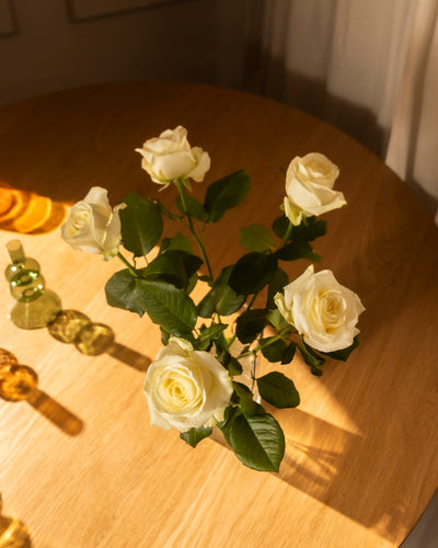 Sechs Weiße Rosen, Symbol für Ehrlichkeit und Respekt, stehen mit grünen Blättern in einer Glasvase auf einem Holztisch im Sonnenlicht. Daneben liegen dekorative Glasobjekte und Orangenscheiben.