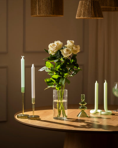 Eine Weiße-Rose-Vase steht auf einem runden Holztisch, umgeben von grünen und weißen Kerzen in verschiedenen Haltern. Warmes Licht schafft ein gemütliches Ambiente, das Respekt und Ehrlichkeit widerspiegelt.