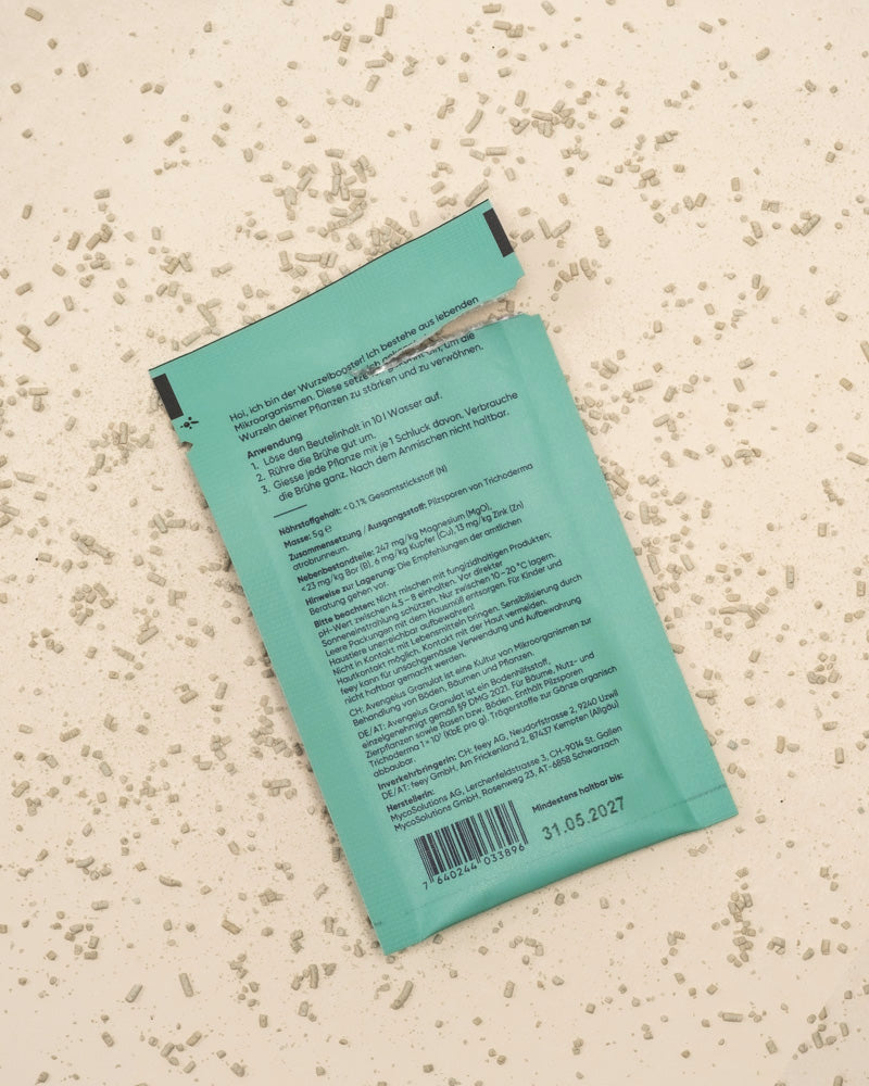 Ein türkisfarbenes Wurzelbooster-Päckchen liegt auf einer leicht gesprenkelten Oberfläche, umgeben von verstreuten weißen Körnchen. Die aufgerissene Packung zeigt auf der Rückseite Text und einen Barcode. Verfallsdatum: 31.05.2027.