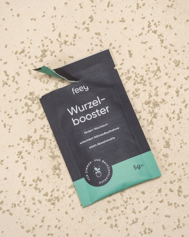 Eine geöffnete Wurzelbooster-Packung liegt auf einer hellen Unterlage, aus der das Granulat herausquillt. Die schwarz-grüne 5g-Packung mit deutschem Text ist zur Förderung des Pflanzenwachstums bestimmt.