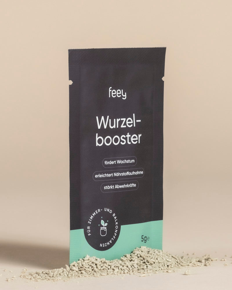 Eine schwarz-grüne 5g-Wurzelbooster-Packung steht aufrecht auf einer beigen Fläche mit leichtem Pulver im Vordergrund. Der deutsche Text hebt die Vorteile für das Pflanzenwachstum hervor und betont, dass das Produkt das Pflanzenwachstum verbessert.