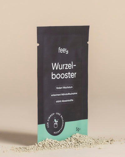 Eine schwarz-grüne 5g-Wurzelbooster-Packung steht aufrecht auf einer beigen Fläche mit leichtem Pulver im Vordergrund. Der deutsche Text hebt die Vorteile für das Pflanzenwachstum hervor und betont, dass das Produkt das Pflanzenwachstum verbessert.