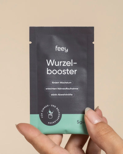 Eine Hand hält eine kleine, blaugrüne und schwarze "Wurzelbooster"-Packung mit deutschem Text, die das Pflanzenwachstum, die Nährstoffaufnahme und die Widerstandsfähigkeit von Zimmer- und Balkonpflanzen fördert. Das 5-g-Produkt enthält Trichoderma atrobunneum.