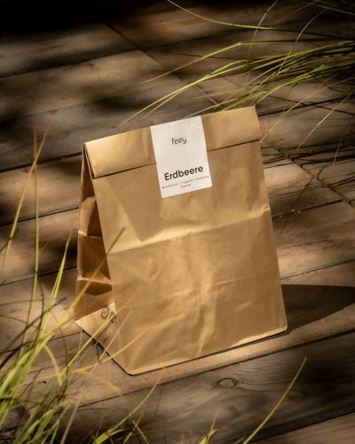 Eine braune Papiertüte mit der Aufschrift „Gartenerdbeere“ der Marke „feey“ liegt auf einem sonnenbeschienenen Holzdeck. Dünne grüne Grashalme rahmen die Tüte ein und deuten auf die bald blühenden, leuchtenden Erdbeeren des Produkts Erdbeere (Wurzelstock) hin.