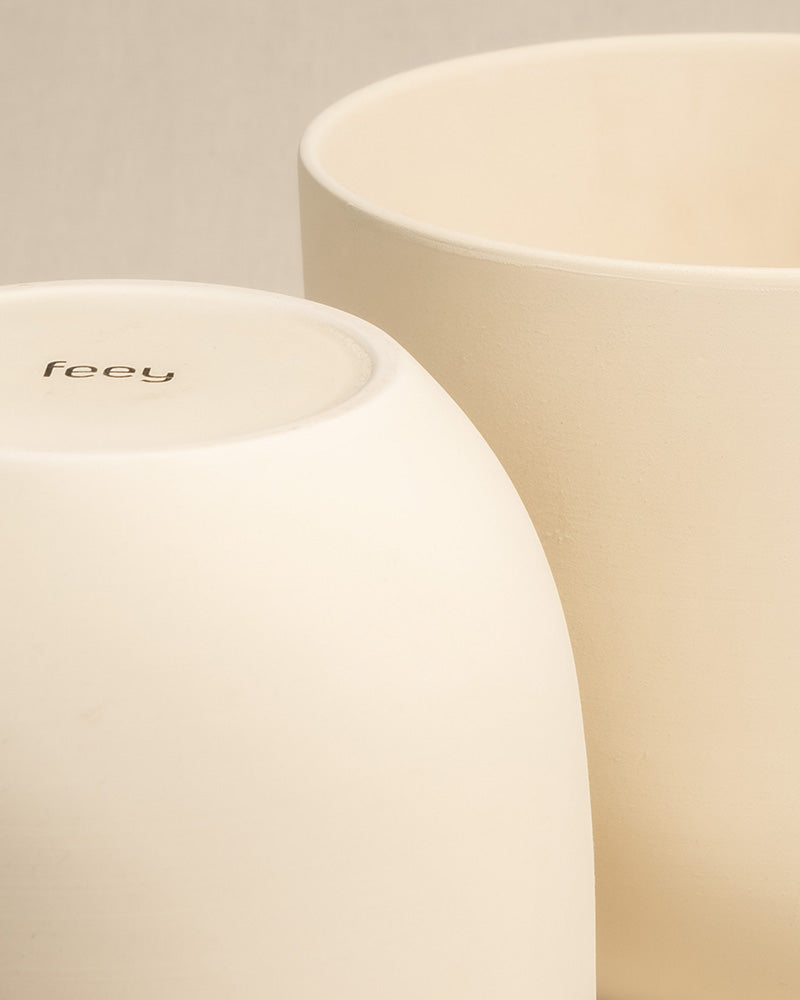 Nahaufnahme von zwei cremefarbenen Keramik-Topfsets 'Direito' (2 × 18, 2 × 14) mit glatten Oberflächen. Ein Topf steht aufrecht, während der andere umgedreht ist, sodass der Markenname „feey“ auf der Unterseite aufgedruckt ist. Der Hintergrund ist ein hellbeiger Stoff, ideal für die Präsentation von Zimmerpflanzen.