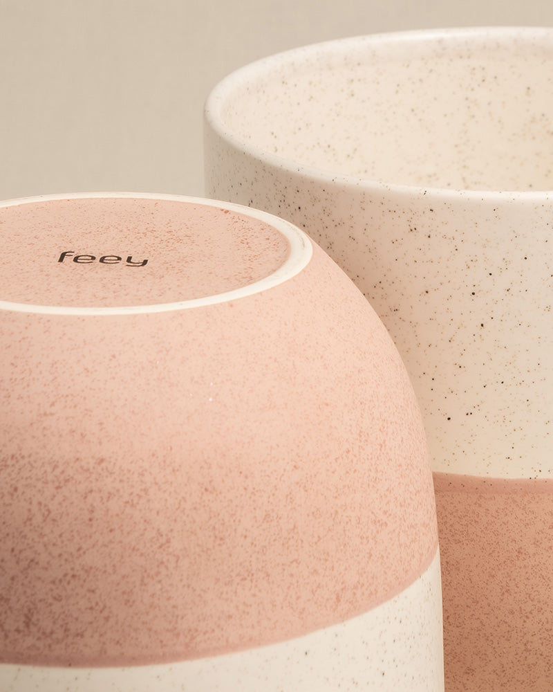 Nahaufnahme von zwei handgefertigten Keramiktassen mit gesprenkeltem Muster. Eine Tasse steht aufrecht und ist in gedecktem Weiß gehalten, während die andere umgedreht ist und ein rosa-weißes Farbschema und den Markennamen „feey“ auf der Unterseite offenbart. Ideal für Minimalisten oder in Kombination mit dem Keramik-Topfset „Variado“ (2 × 16, 2 × 14), um jedem Raum Charme zu verleihen.