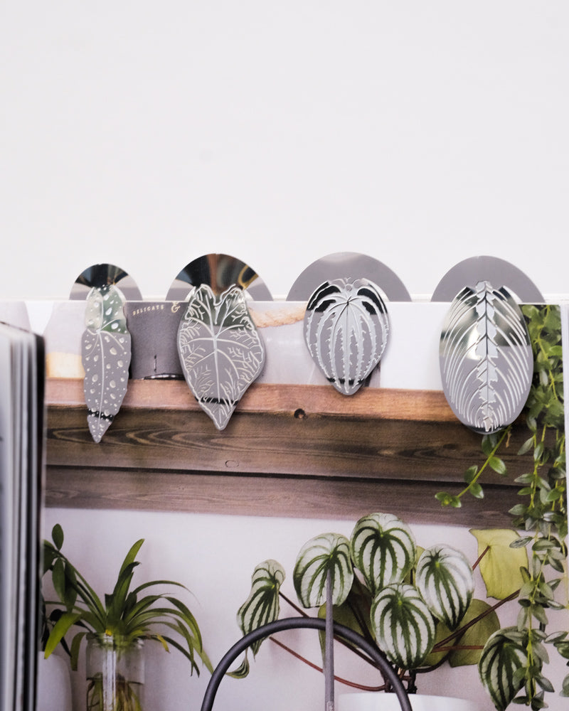 Vier Buchzeichen: Zimmerpflanzen-Blätter aus Metall in Form verschiedener Blätter sind oben auf einem Buch befestigt. Jedes Lesezeichen weist komplizierte Blattmuster auf. Unter den Lesezeichen zeigt das Buch das Bild eines Holzregals, das mit grünen Topfpflanzen geschmückt ist.