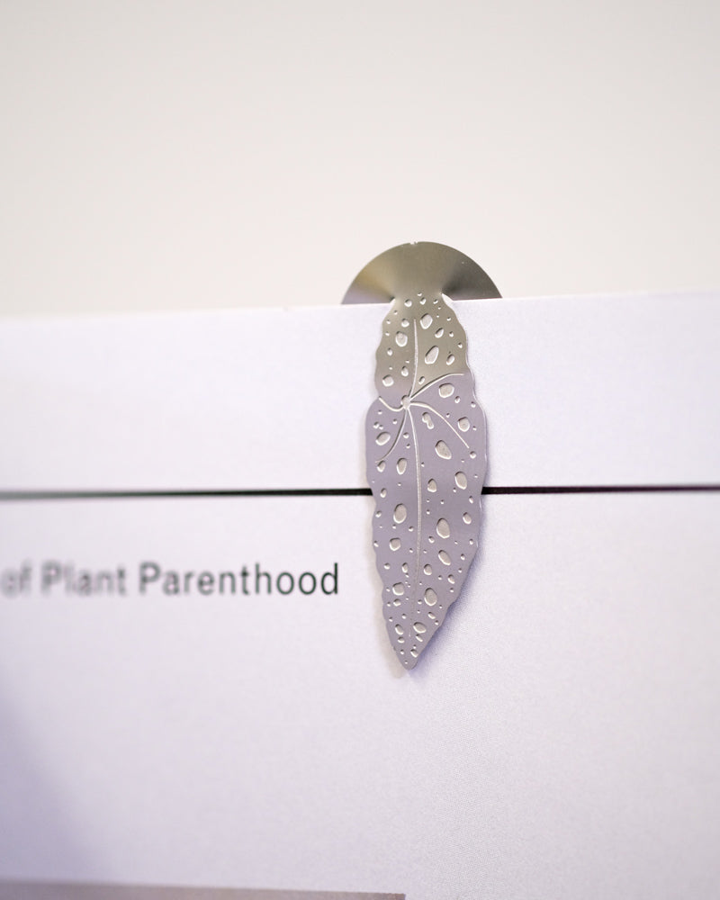 Eine Nahaufnahme eines metallenen Buchzeichens: Zimmerpflanzen-Blätter in Blattform, teilweise in ein weißes Heft eingelegt. Das Lesezeichen ist mit aufwendigen ausgeschnittenen Mustern sorgfältig gearbeitet und ähnelt Pflanzenblättern mit zahlreichen Löchern. Der sichtbare Text auf dem Heft lautet „Plant Parenthood“.