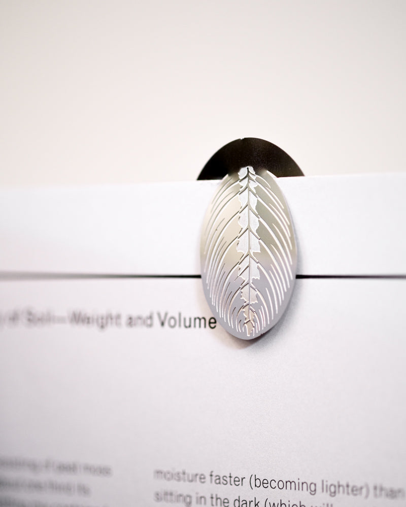 Eine Nahaufnahme eines weißen Papiers, das von einer dekorativen silbernen, blattförmigen Klammer vor einem weißen Hintergrund gehalten wird. Die Büroklammer, die gleichzeitig als Buchzeichen dient: Zimmerpflanzen-Blätter, weist komplizierte ausgeschnittene Details auf. Ein Teil des Textes auf dem Papier ist sichtbar, aber teilweise unscharf.