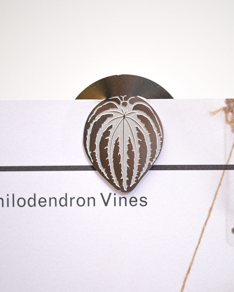 Eine Nahaufnahme eines metallischen Pflanzenlesezeichens in Form eines Blattes mit aufwendigen Ausschnitten, das am Rand einer Seite befestigt ist. Auf der Seite ist teilweise der Text „Philodendron Vines“ sichtbar. Der weiße Hintergrund hebt das filigrane Design des Buchzeichens: Zimmerpflanzen-Blätter hervor.