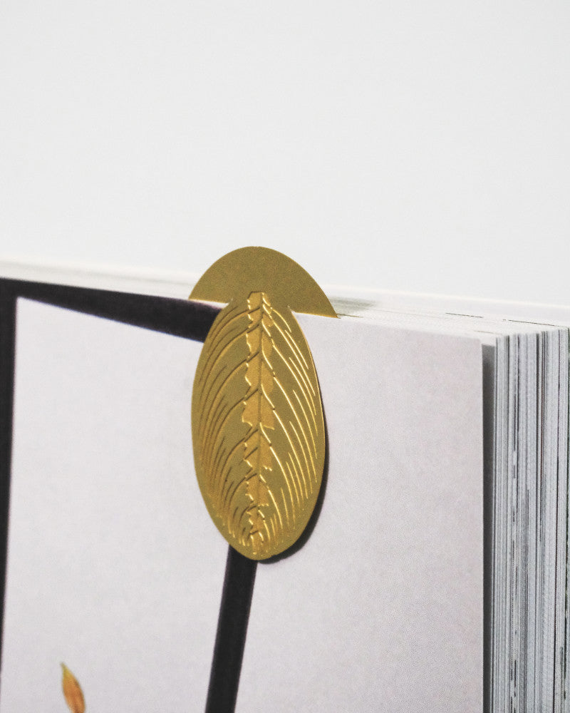 Ein goldenes, blattförmiges Metallbuchzeichen: Zimmerpflanzen-Blätter ist oben an einem offenen Buch befestigt. Das Lesezeichen weist komplizierte Aderndetails auf den Pflanzenblättern auf. Die Seiten des Buches scheinen weiß zu sein, mit einigen undeutlichen, farbigen Details, die an der Seite sichtbar sind. Der Hintergrund ist schlicht weiß.