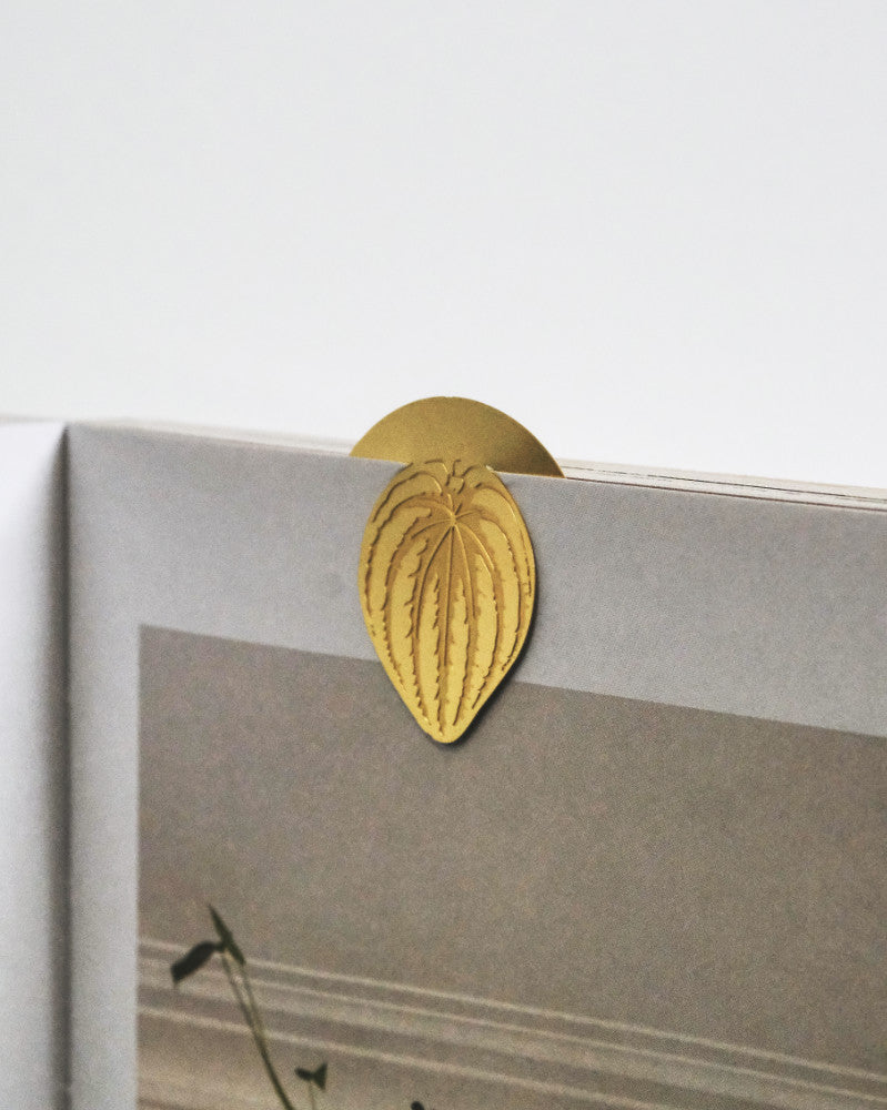 Eine Nahaufnahme eines Buchzeichens aus Goldmetall: Zimmerpflanzen-Blätter in Blattform, das in der oberen Ecke eines Buches angebracht ist. Das Lesezeichen mit detaillierten Blattadern ragt über die Seiten hinaus, während ein Teil einer Seite mit Text und Bildern zu sehen ist.