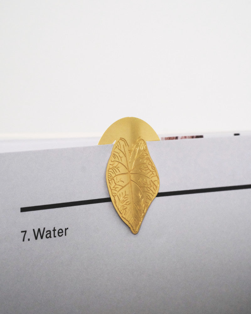 Ein goldmetallisches, blattförmiges Buchzeichen: Zimmerpflanzen-Blätter, das an der oberen Ecke einer Seite mit der Überschrift „7. Wasser“ angebracht ist. Die detailreichen Pflanzenblätter mit Adern sind über den Rand der Seite gesteckt. Der Hintergrund ist schlicht und weiß, was seine Eleganz und korrosionsbeständige Qualität unterstreicht.