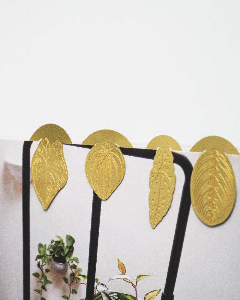 Vier goldmetallische Buchzeichen: Zimmerpflanzen-Blätter, jedes in Form eines anderen Pflanzenblatts, sind oben an einem weißen Buch oder Notizbuch befestigt. Im Hintergrund ist teilweise etwas Grün zu sehen, was der Szene einen Hauch von Natur verleiht.