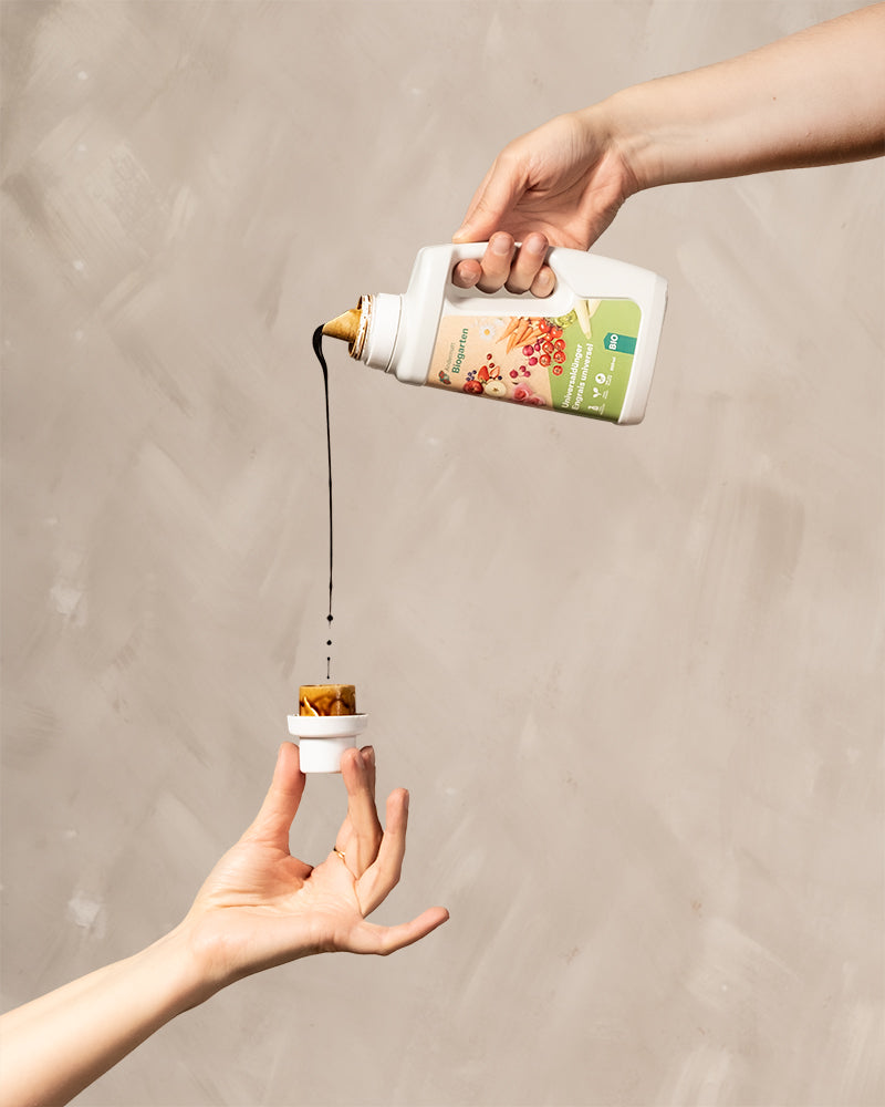 Eine Hand hält die Flüssigdüngerflasche und giesst Dünger in den Flaschenkappe, die von einer 2. Hand gehalten wird.