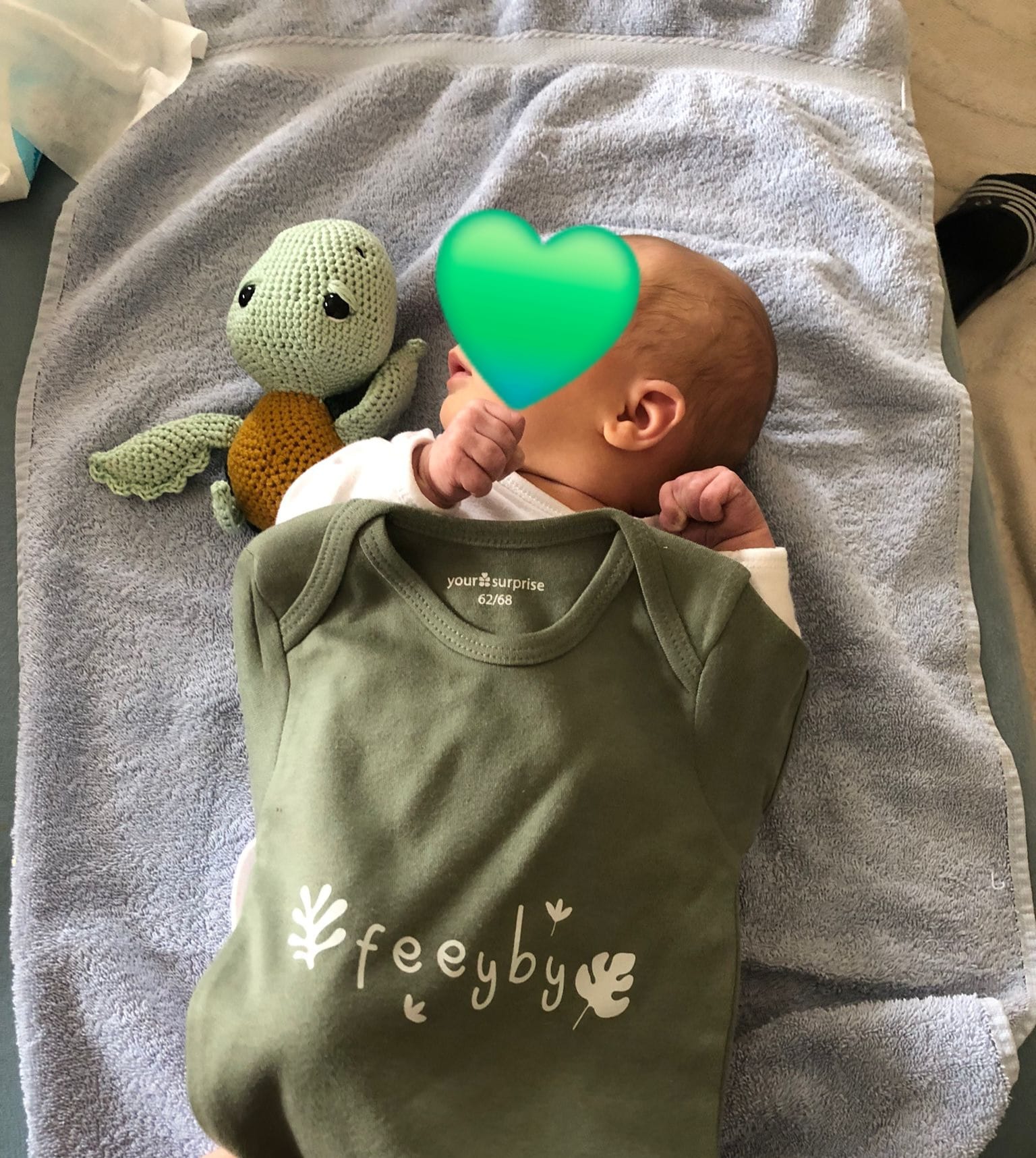 Ein mit Herz-Emoji zensiertes Baby liegt neben einer gehäkelten Schildkröte und einem Strampelanzug mit Aufschrift «feeyby».