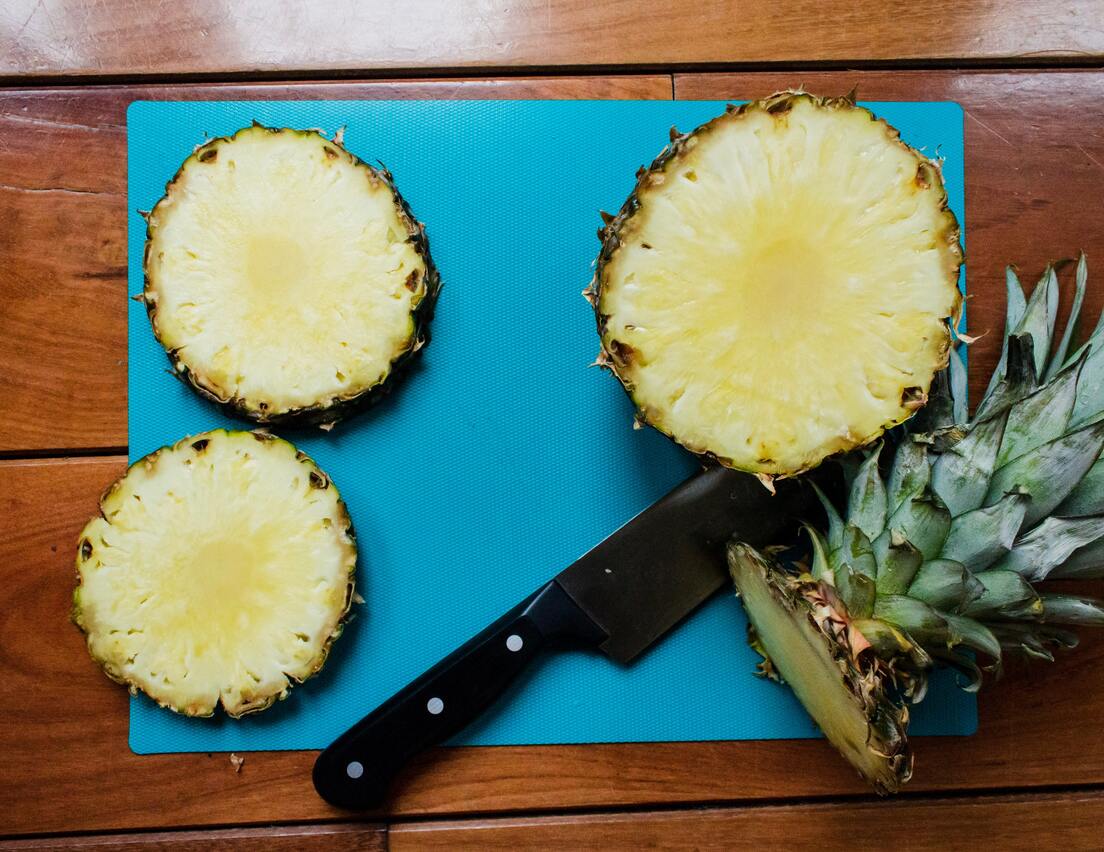 Ein schwarzes Messer, drei Scheiben Ananas und der abgeschnittene Schopf auf einem blauen Schneidebrett