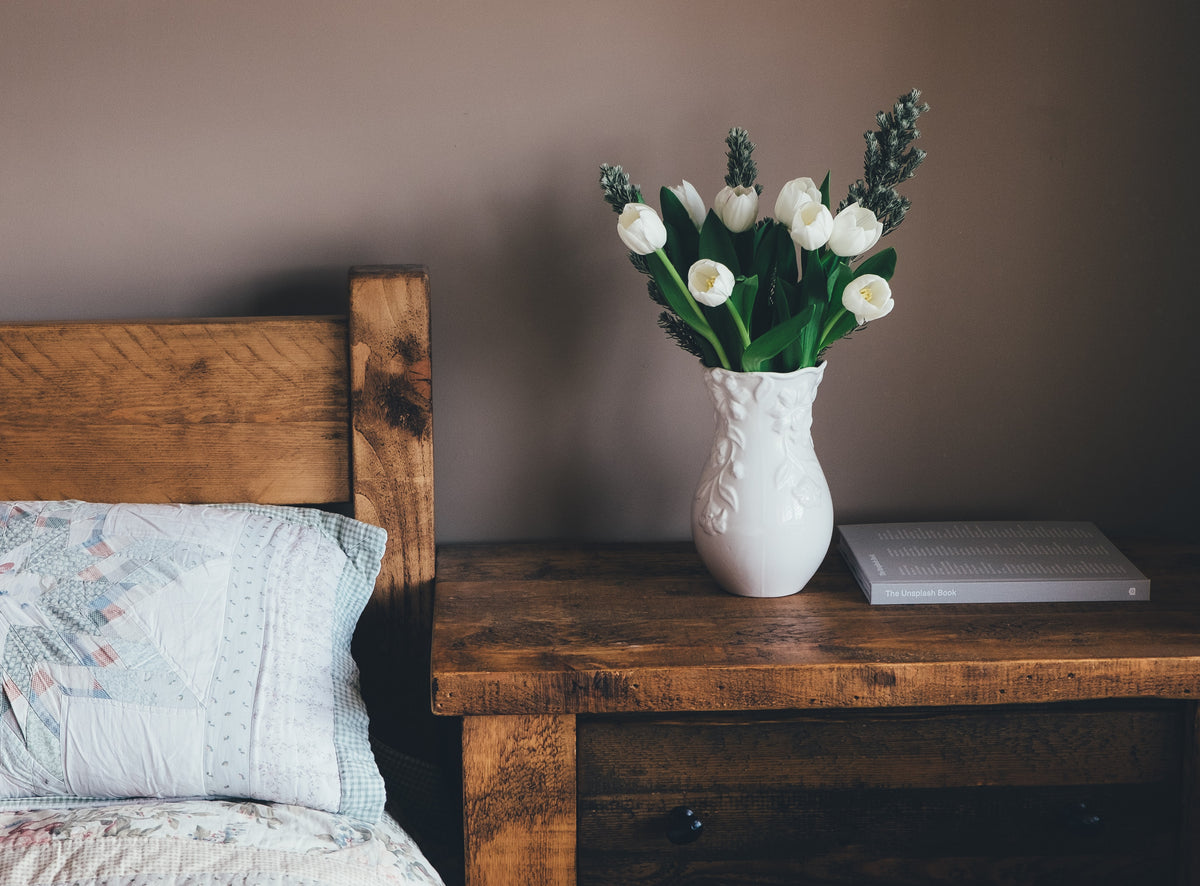 Ein hölzerner Nachttisch steht neben einem hölzernen Bett vor einer dunklen Wand. Auf dem Nachttisch steht eine weisse Vase mit weissen Tulpen darin.