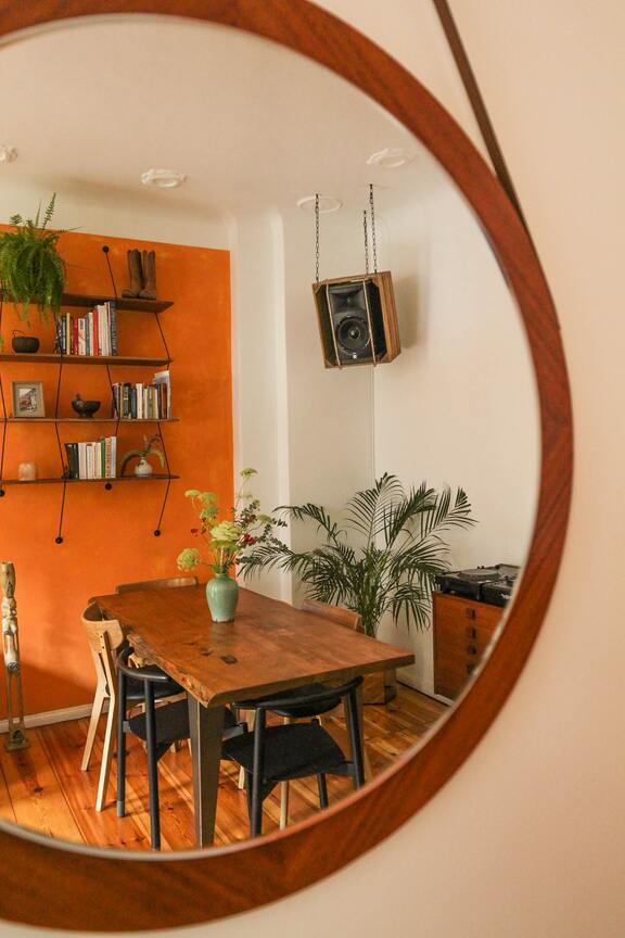 Bild eines runden Spiegels, darin sieht man einen Holztisch, einen Holzboden, eine orangene Wand mit Wandregal und Streifenfarn auf der obersten Etage, in der Ecke eine Goldfruchtpalme und auf dem Tisch gelbe Trockenblumen