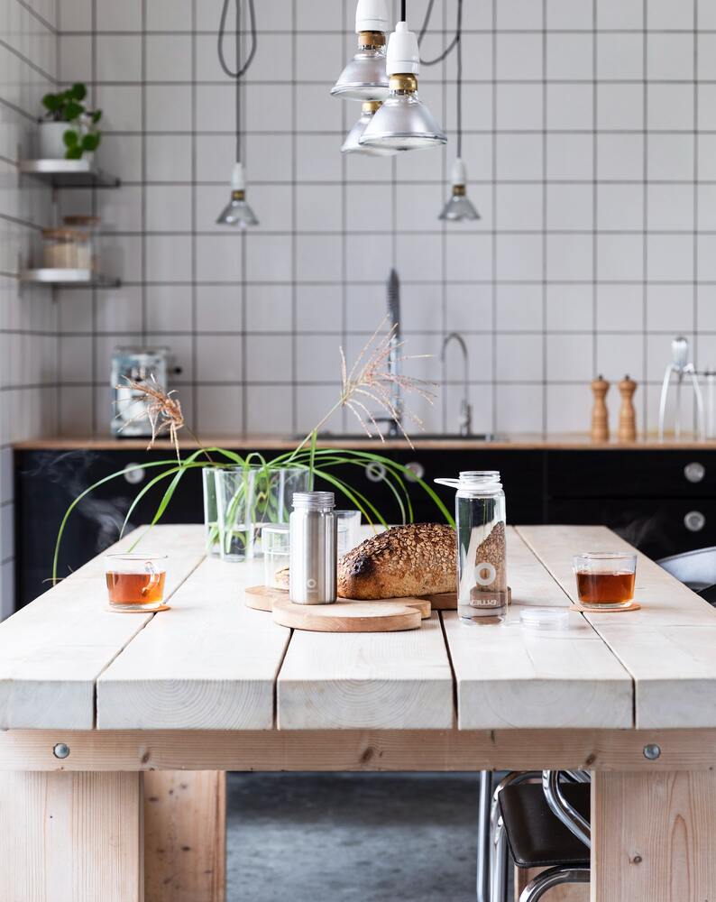 Grünlilie in durchsichtigem Gefäss auf einem rustikalen Holztisch, dahinter eine quadratisch weiss gekachelte Küchenwand