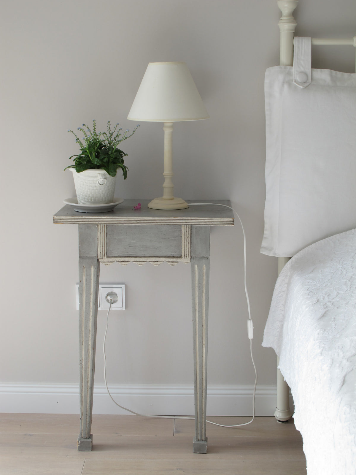 Auf einem grauen Nachttisch steht eine weisse Lampe und eine kleine Pflanze in einem weissen Topf. Daneben steht ein weisses Bett mit weisser Bettwäsche. 