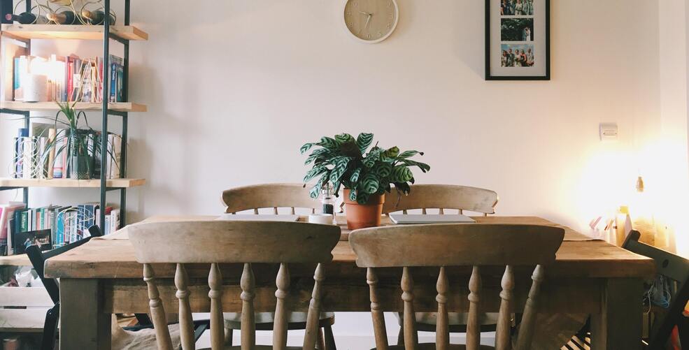 holziger Esstisch mit vier Holzstühlen, dahinter eine Uhr und ein Bild an der Wand sowie ein Bücherregal mit diversen Pflanzen, auf dem Tisch selbst eine Calathea zentral als Blickfang
