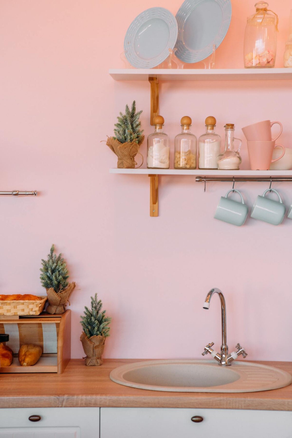 Rosa Pastellwand, davor eine hölzerne Kücheninsel  mit rundem lavabo, dahinter ein dezentes Wandregal mit Lebensmitteln in Glasbehältern und rosa Tassen, dazwischen kleine Pflanzen
