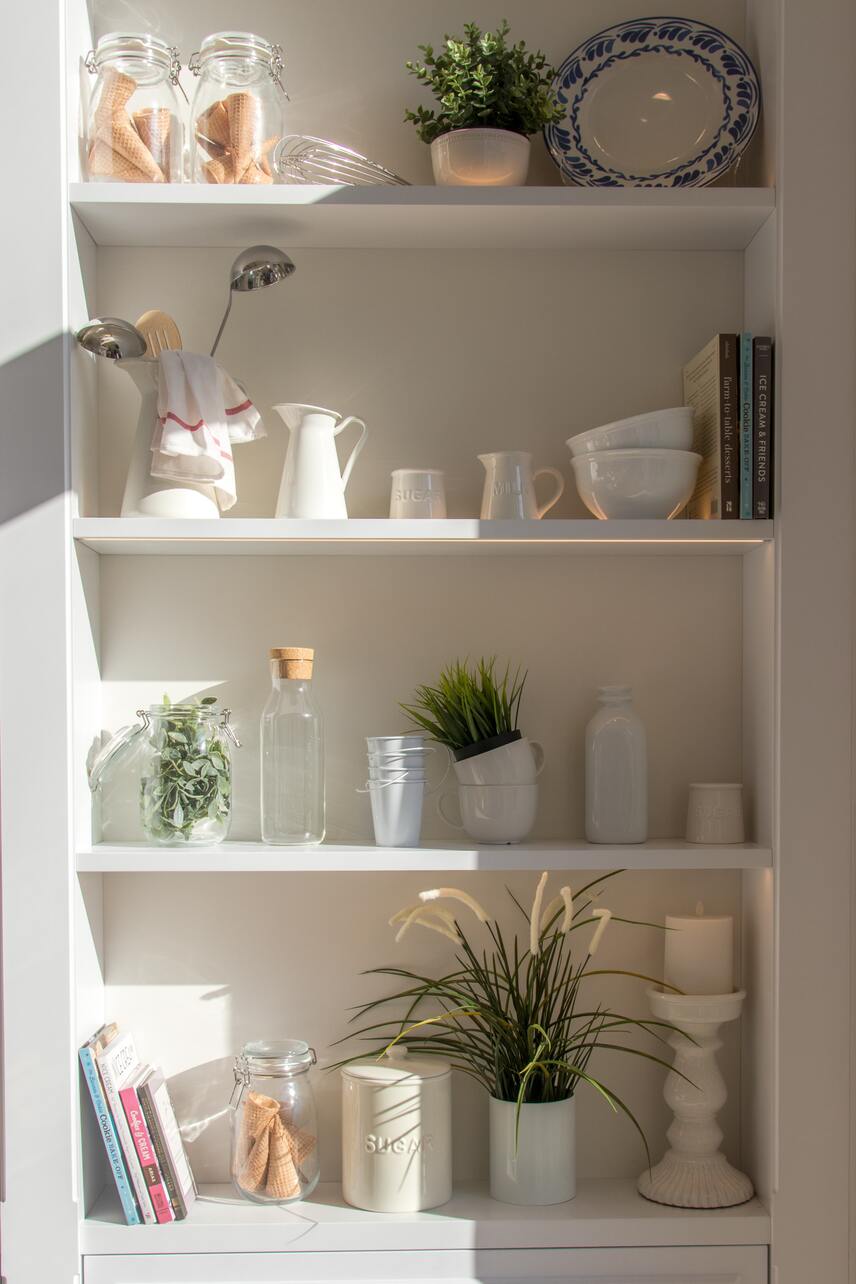 Weisses, geschlossenes Küchenregal mit weissem Geschirr, Pflanzen in weissen Übertöpfen und Lebensmitteln in Glascontainern