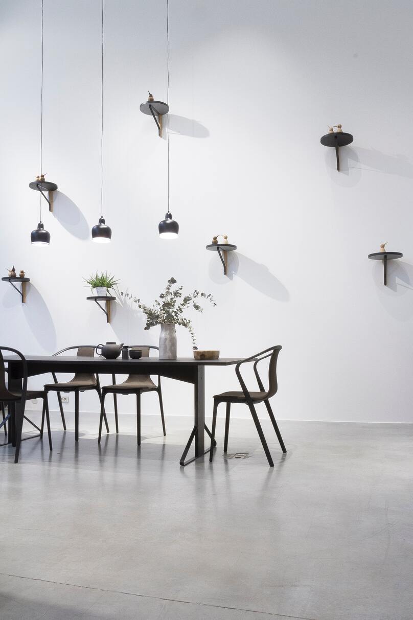 Langer, dunkler Tisch, schlichte Stühle, eine weisse Wand mit vielen minimalistischen Regalen, darauf kleine Pflanzen und auf dem Tisch ein Strauss Trockenblumen
