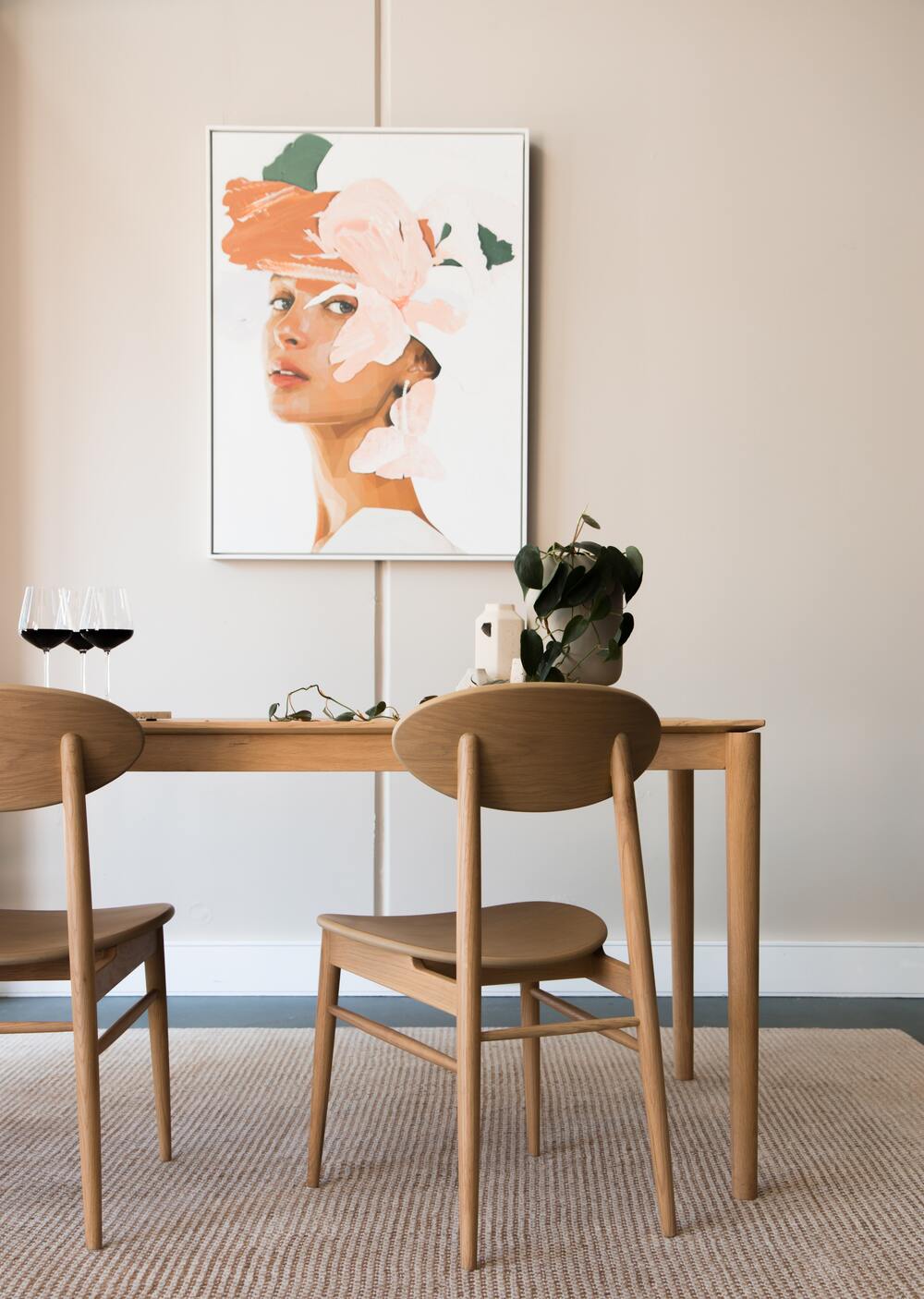Holztisch mit Weingläsern und Philodendron auf dem Tisch, davor zwei Holzstühle und dahinter an der Wand ein Artprint eines Frauenkopfs