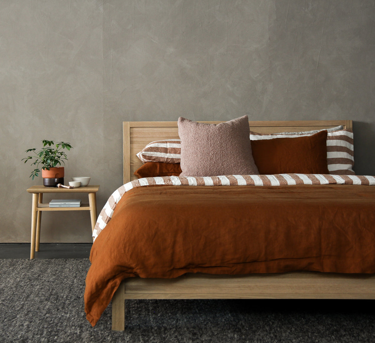Ein Bett steht vor einer grauen Wand. Die Bettdecke ist kufperfarbig. Es hat mehrere Kissen. Das Bett ist aus Holz. Der Nachttisch ist auch aus Holz. Auf dem Nachttisch steht eine Pflanze, eine Schefflera.