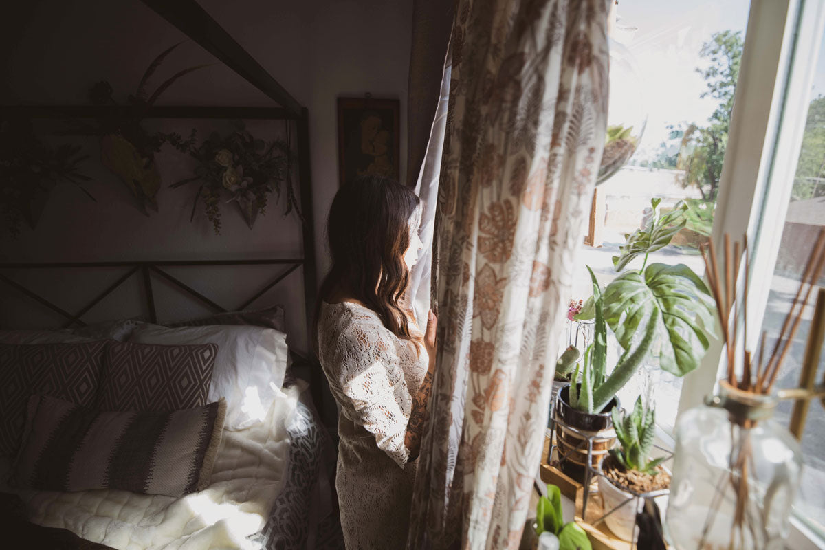 Abgedunkeltes Schlafzimmer, eine Frau öffnet die Vorhänge, eine Monstera und andere Pflanzen auf dem Fensterbrett