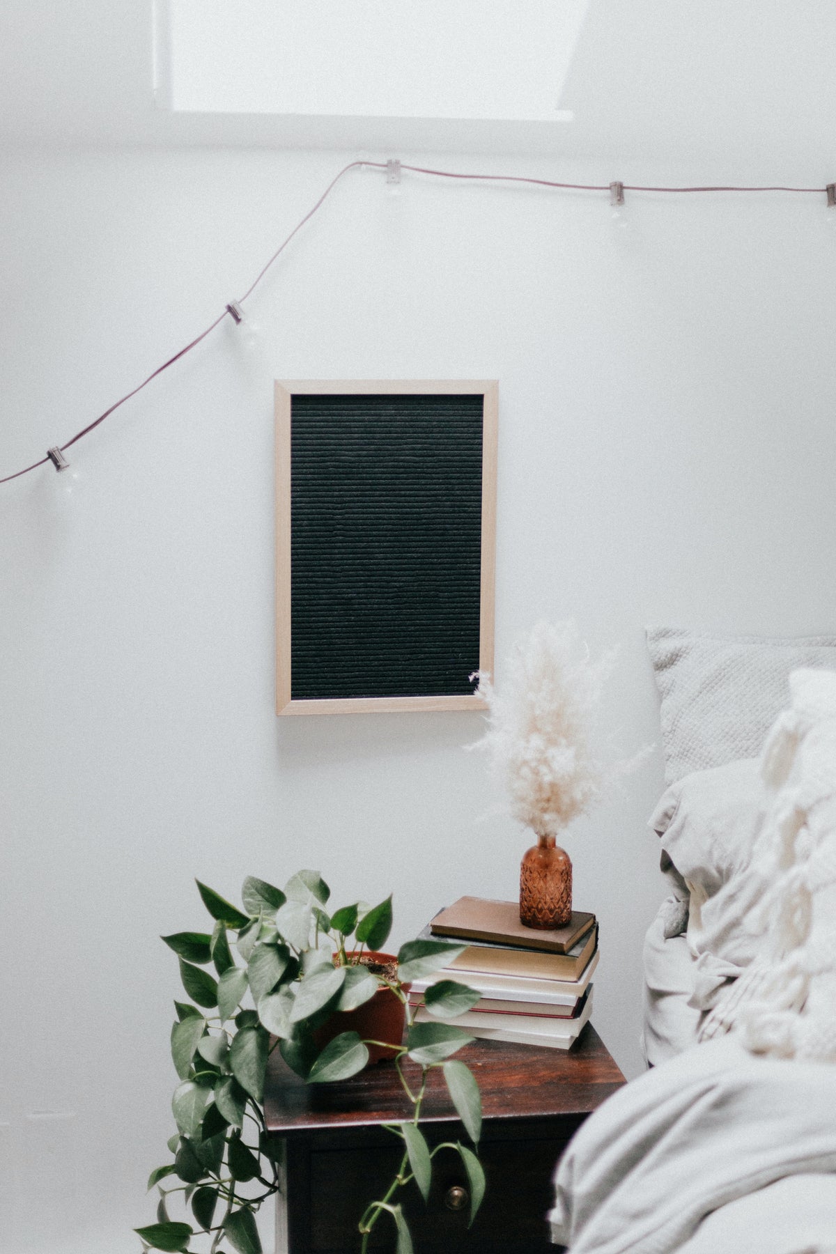 Ein Nachttisch aus dunklem Holz steht neben einem Bett mit weisser Bettwäsche. An der Wand hängt ein schwarzes Bild und eine Lichterkette. Auf dem Nachttisch hat es eine Efeutute, Bücher und eine Vase mit weissen Blumen.