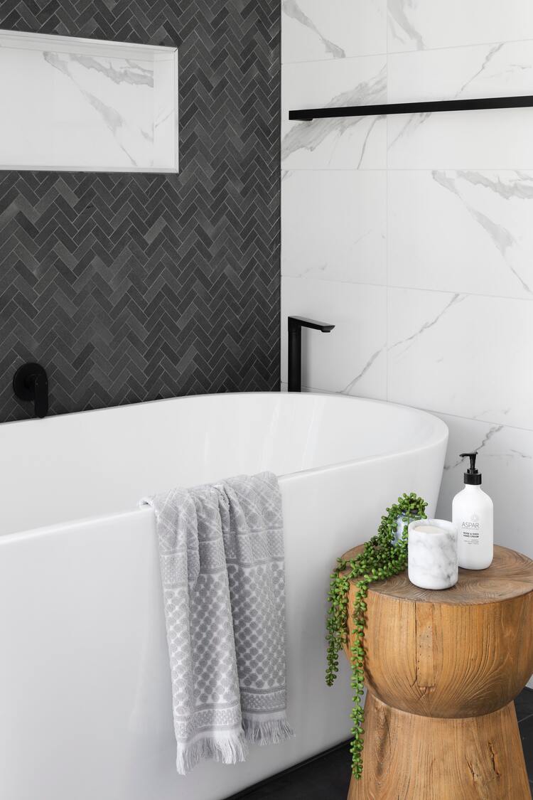 Ovale Badewanne, je eine grau und eine marmoriert gemusterte Wand, vor der Wanne ein runder Holz-Beistelltisch mit Seife und einer kleinen Hängepflanze