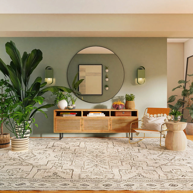 Grosse buschige Zimmerpflanzen auf einem weiss gemusterten Teppich rahmen ein flaches Sideboard und einen grossen, runden Spiegel darüber ein