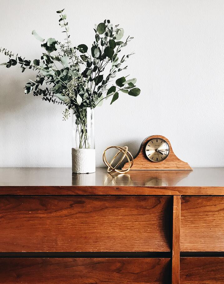 Blassgrünes Trockenblumen-Arrangement in einer durchsichtigen Vase neben einer altertümlichen Uhr und einem goldenen Dekoelement auf einem braunen Möbel