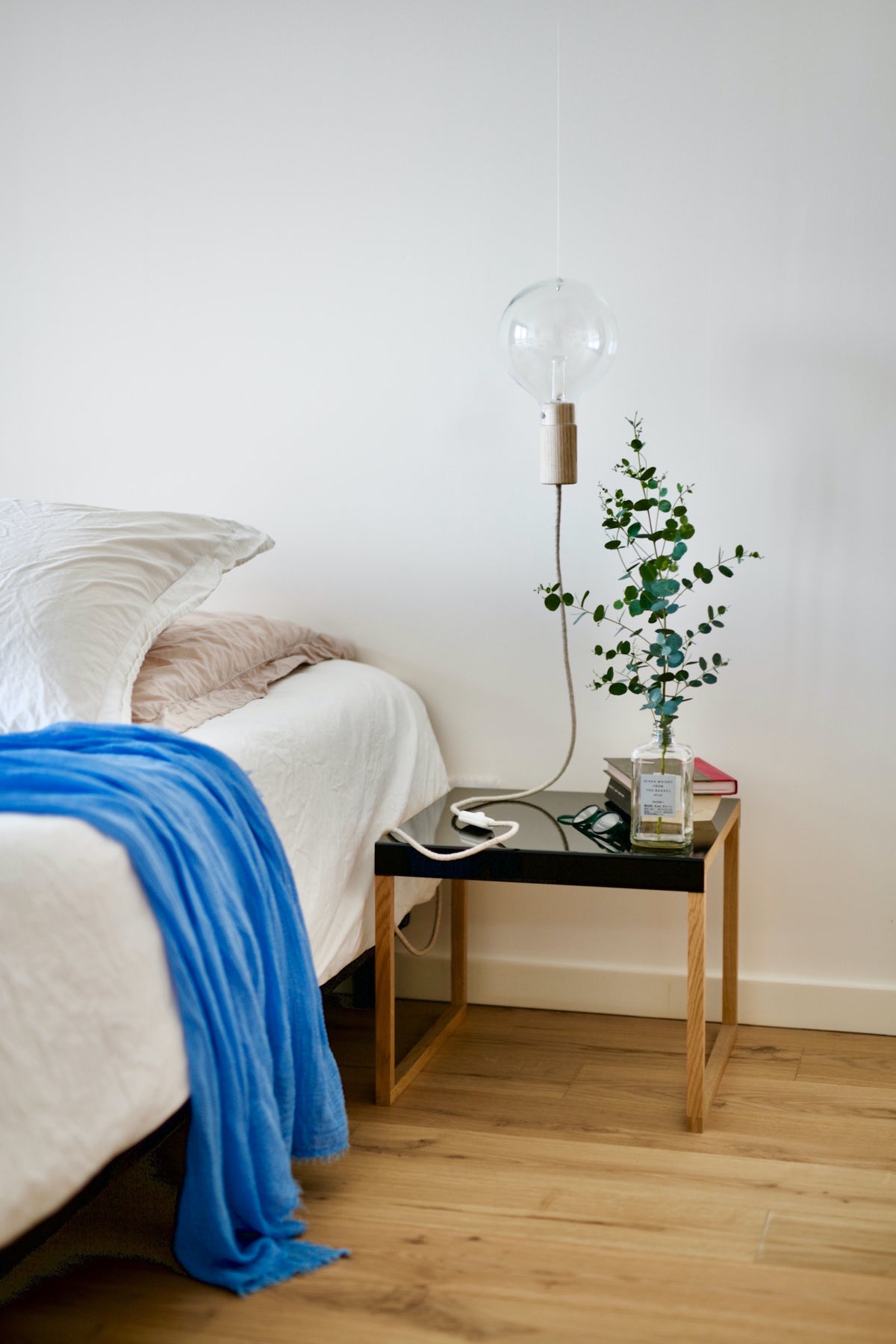 Ein schwarzer minimalistischer Nachttisch steht vor einer weissen Wand neben einem Bett mit weisser Bettwäsche. Auf dem Nachttisch hat es eine Lampe, Bücher, eine Lesebrille und eine Vase mit einem Ast.