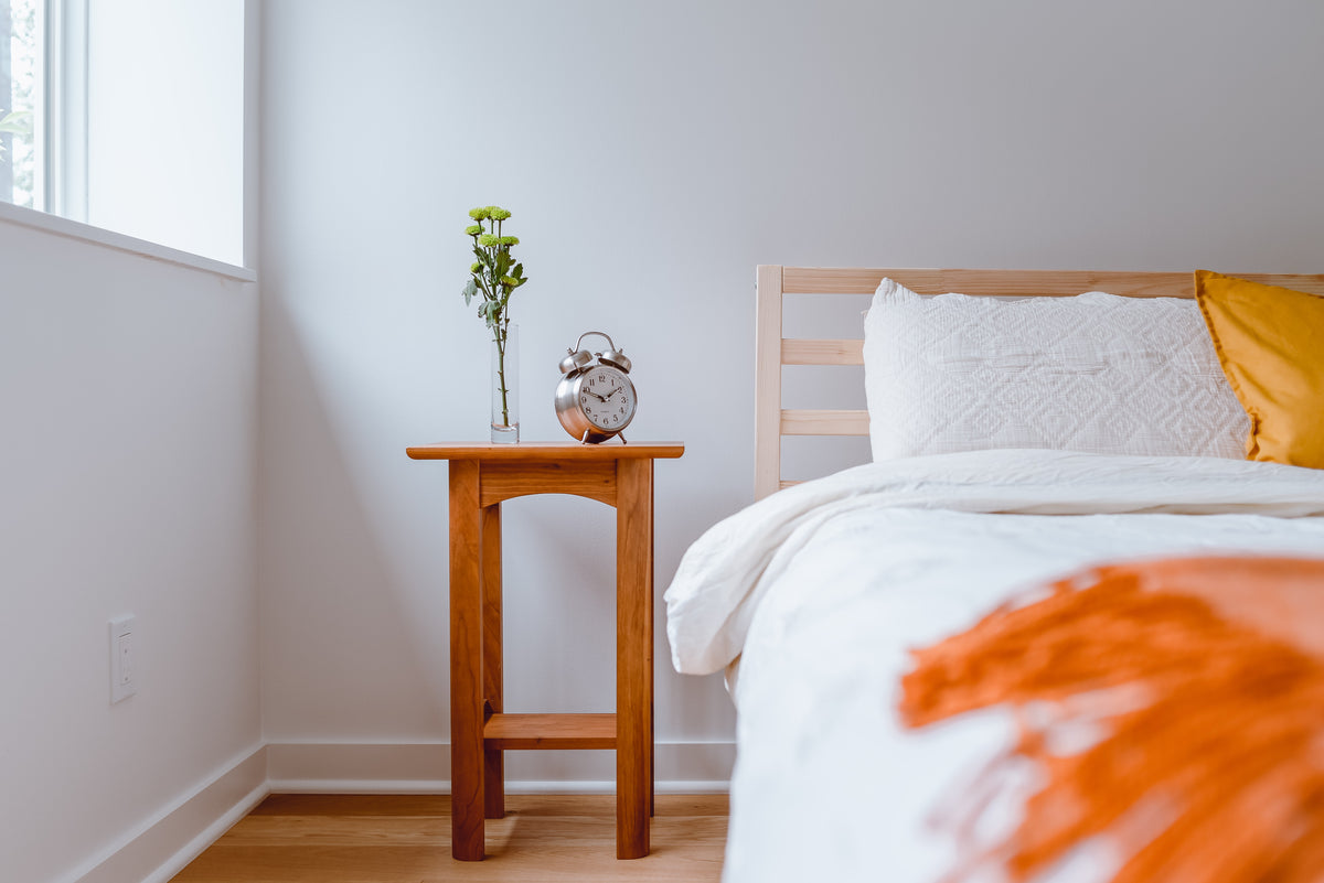 Ein hölzernen Nachttisch steht neben einem hölzernen Bett mit weisser und oranger Bettwäsche. Auf dem Nachttisch steht ein Wecker und eine Vase mit Blumen drin.