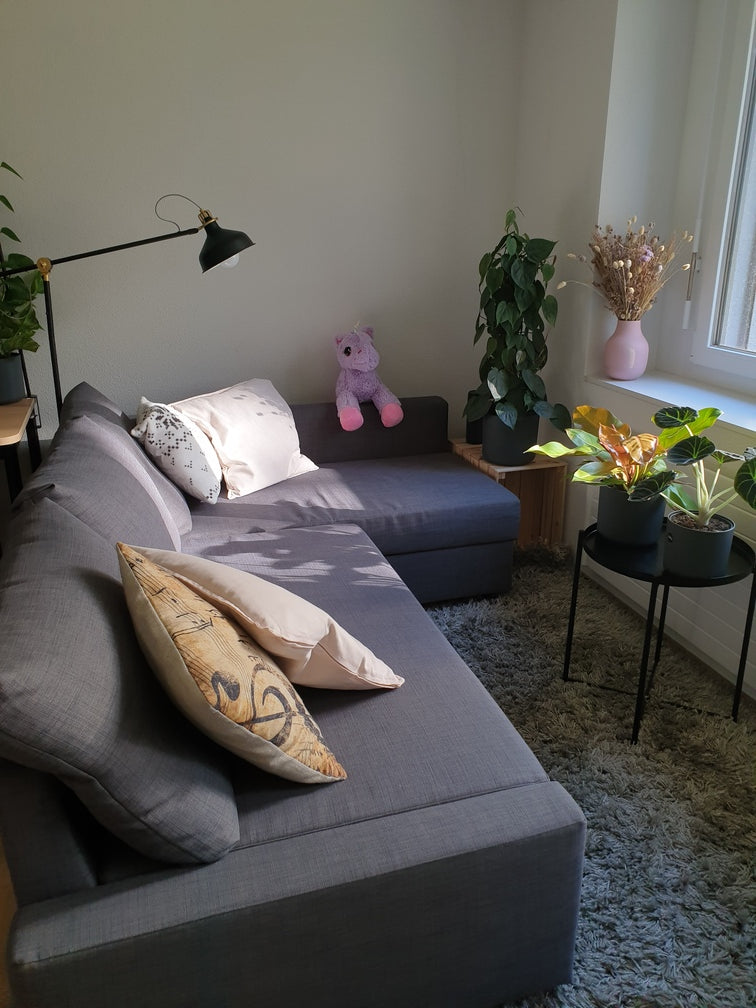 Graues Sofa mit beigen Kissen auf einem grauen, flauschigen Teppich, davor verschiedene Pflanzen auf Tischchen und Holzkiste sowie einem Trockenblumen-Strauss auf dem Fensterbrett
